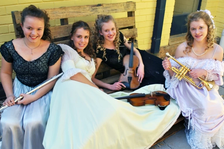 Four students in period costumes, holding musical instruments including a flute, violins and a trumpet.