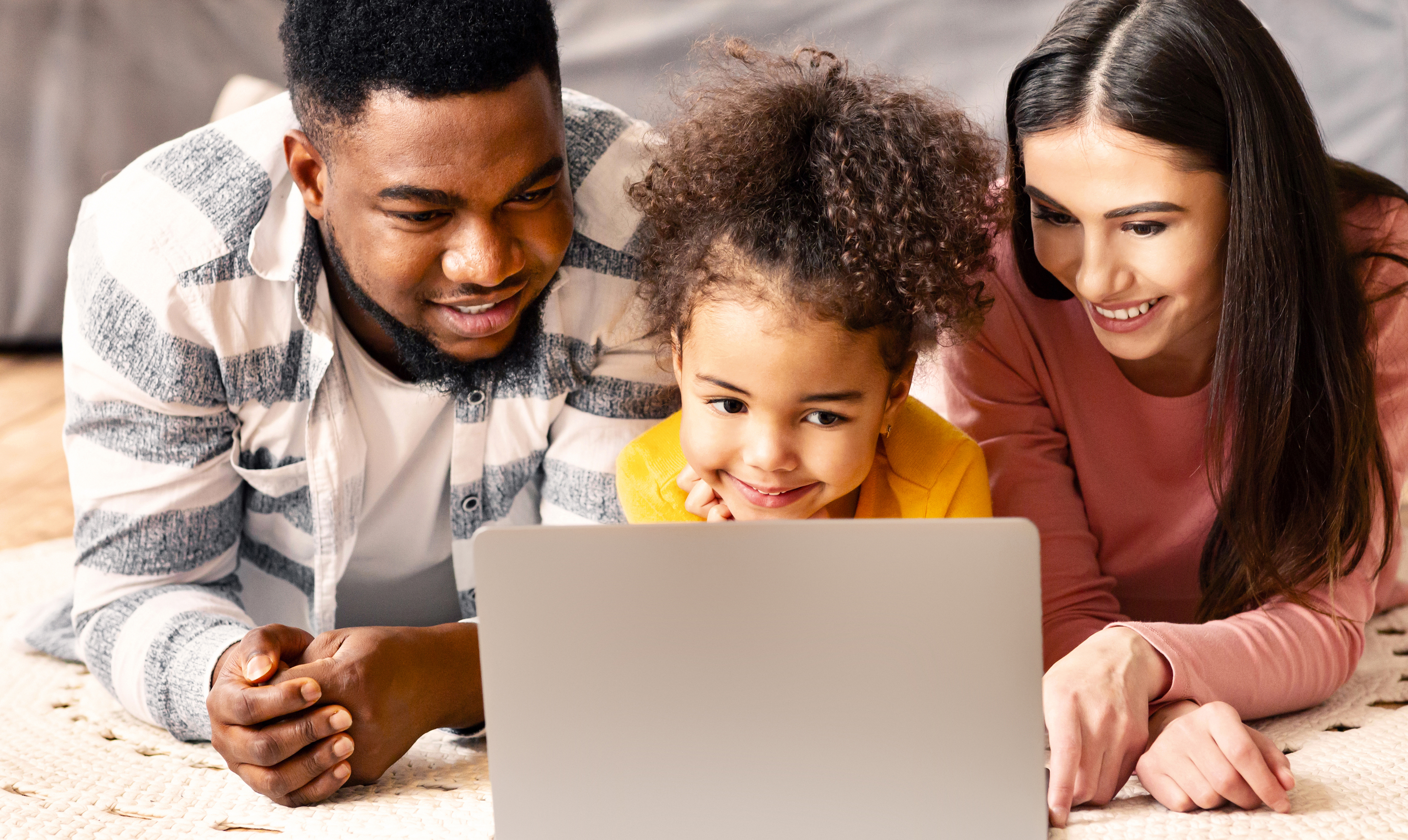 A happy, diverse family with a young child lies on the floor, smiling and looking together at a laptop screen, representing supported family learning and togetherness.