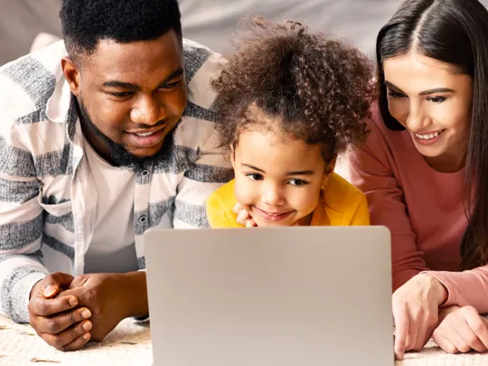 A happy, diverse family with a young child lies on the floor, smiling and looking together at a laptop screen, representing supported family learning and togetherness.