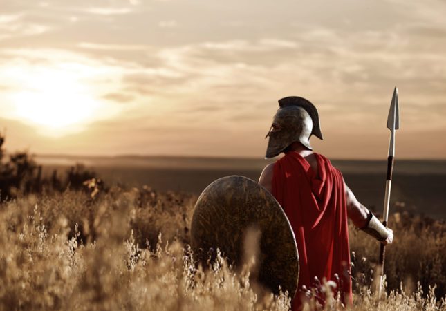 Person dressed as a Roman soldier with spear and shield, looking towards a dramatic sunset over a field