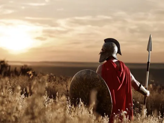 Person dressed as a Roman soldier with spear and shield, looking towards a dramatic sunset over a field