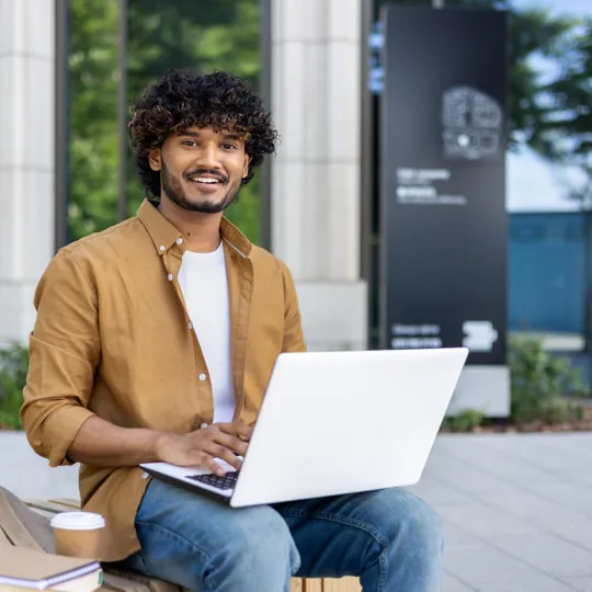 a young man smiling while working on a white laptop and sitting on a wooden bench outside a modern building.