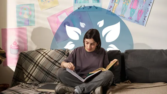 A young person with dark hair sits cross-legged on a patterned couch, engrossed in reading an open book. Various posters, including