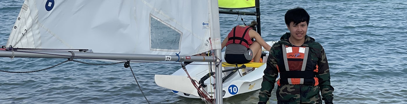 A young person in a life jacket standing by a small sailboat on the water, smiling at the camera.