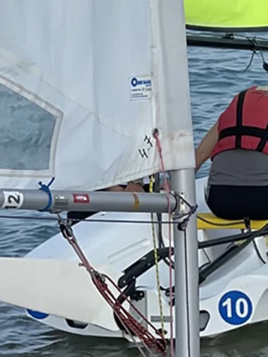 A young person in a life jacket standing by a small sailboat on the water, smiling at the camera.