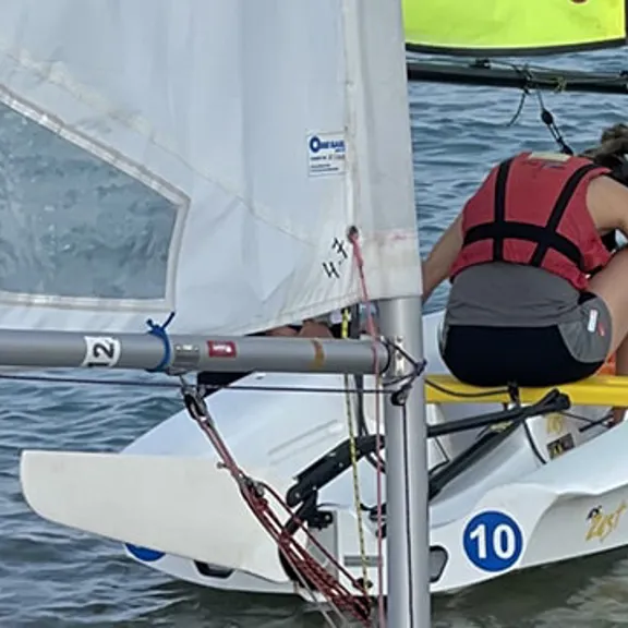 A young person in a life jacket standing by a small sailboat on the water, smiling at the camera.