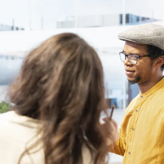 A man wearing a yellow shirt, glasses, and a flat cap is talking seriously with an unidentifiable person in a bright, modern office or study space.