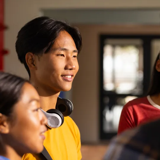 A smiling male student in a yellow shirt and headphones around his neck, engaged in a group conversation with other students on a school campus.