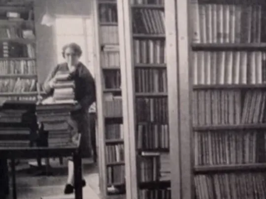 An old black and white photograph of a female library worker in a library. The lady is carrying a big pile of books and walking towards the camera.