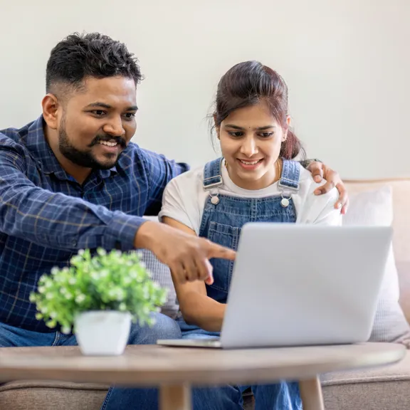 Smiling couple sitting on a sofa looking at a laptop together, with the man pointing at the screen.