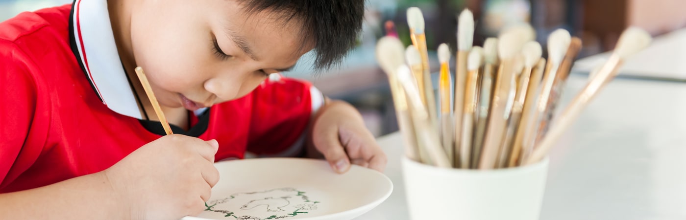 Student focused on painting a ceramic plate, with a pot of paintbrushes nearby on the table