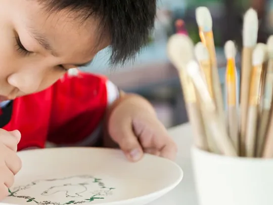Student focused on painting a ceramic plate, with a pot of paintbrushes nearby on the table