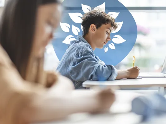 A young student with curly hair is focused on writing during an exam or study session, with a laptop nearby.