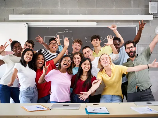 A large, diverse group of students and a teacher cheering and raising their hands in a classroom, with a diagram on the blackboard behind them.