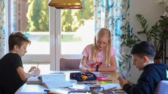 Three children sit together at a dining room table, focused on their books and writing, illustrating a timeless homeschooling environment.