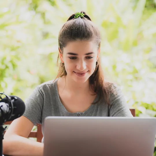 A young woman with a ponytail smiles while working on a laptop with a professional microphone at an outdoor desk in a lush, green setting, representing online learning.