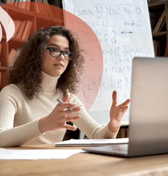 A specialist online tutor gestures towards her laptop as she teaches a complex maths lesson from her study.