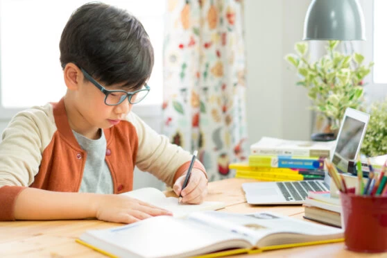 A young boy with short black hair and glasses studying at his bedroom desk.