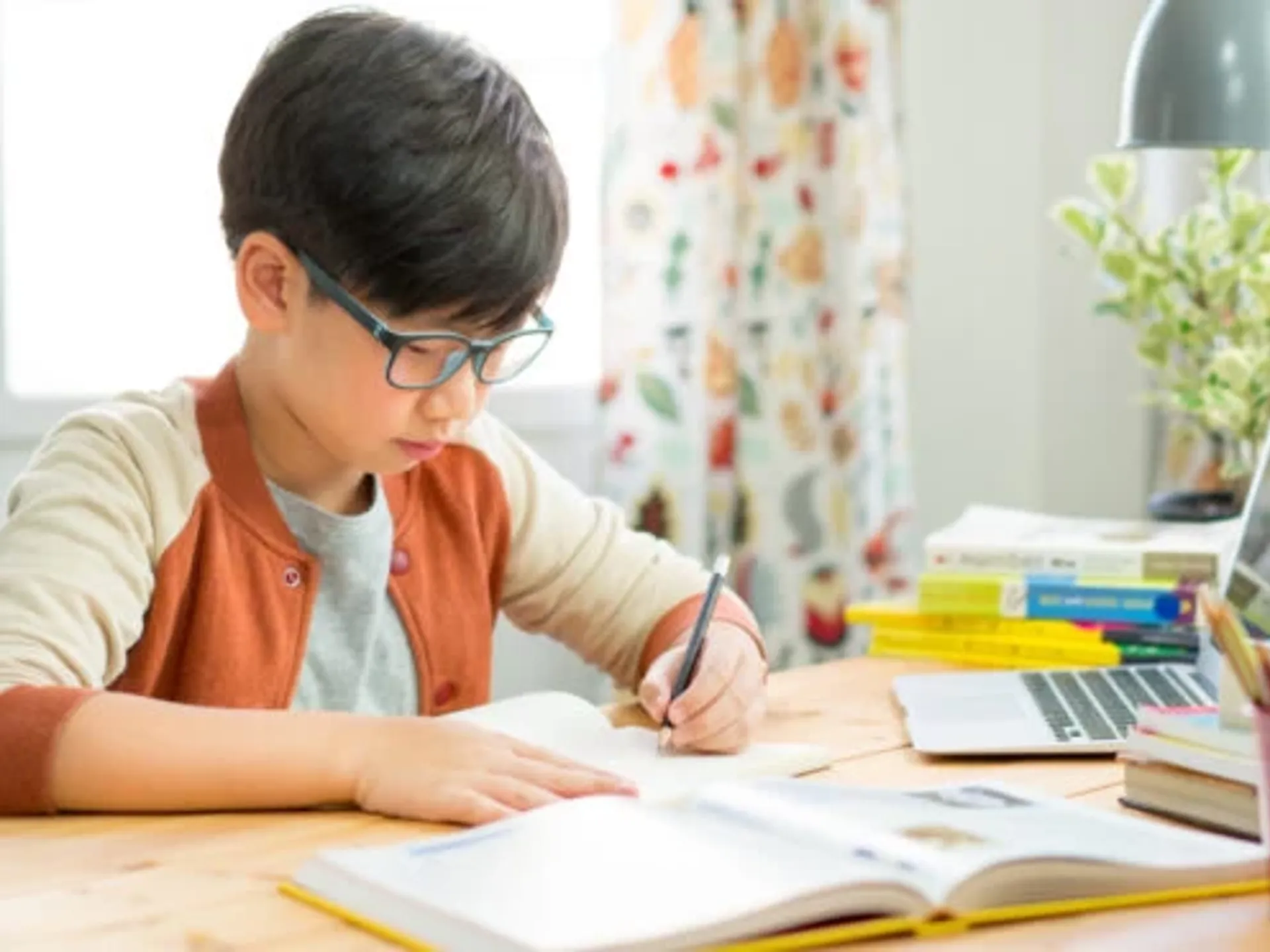 A young boy with short black hair and glasses studying at his bedroom desk.