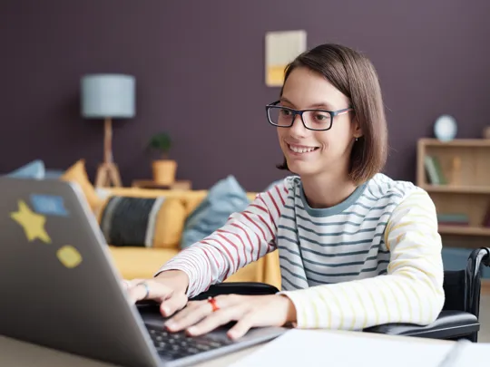 Teenage girl studying on laptop in wheelchair in comfortable home setting