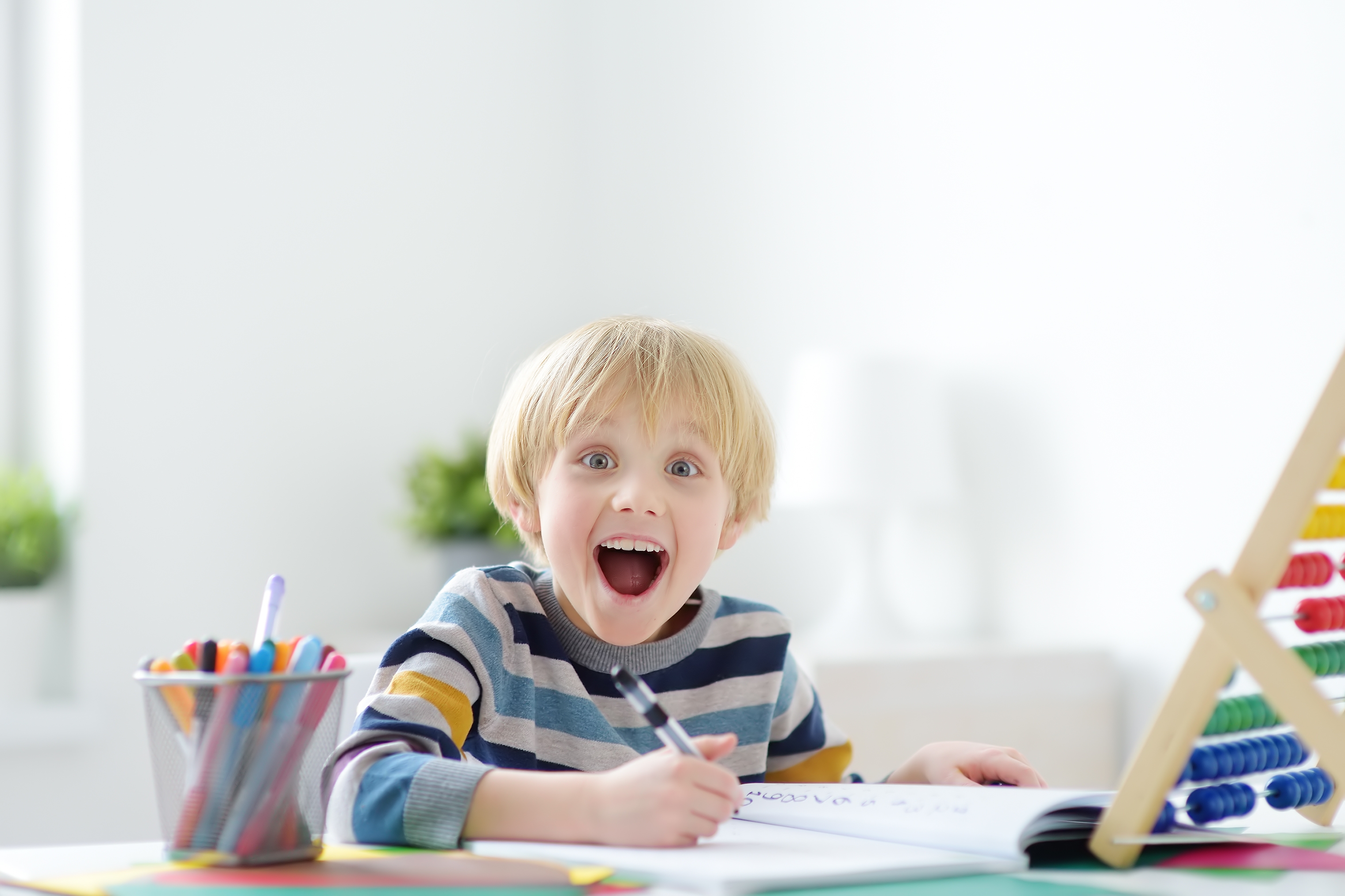 A young student is focused on writing or drawing in a notebook with a pen at a bright white desk. To his left is a cup of colorful pens and to his right is a large wooden abacus.