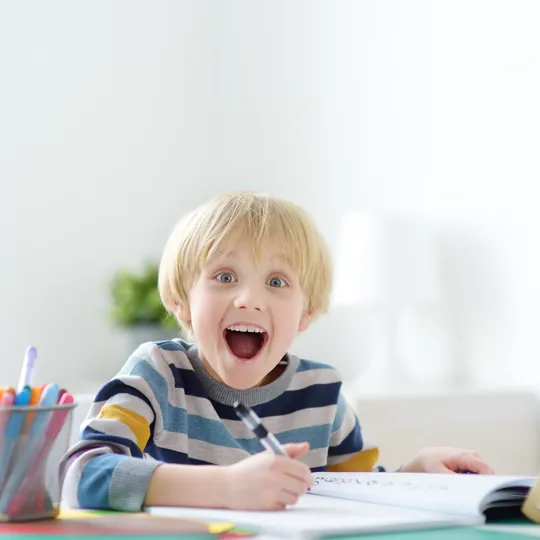 A young student is focused on writing or drawing in a notebook with a pen at a bright white desk. To his left is a cup of colorful pens and to his right is a large wooden abacus.