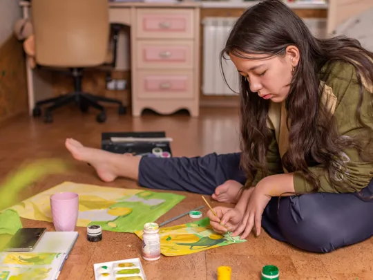 Teen girl painting a colourful artwork on the floor of her bedroom, surrounded by art supplies.