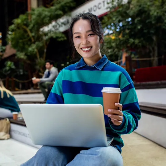 A young woman in a striped sweatshirt smiles while working on a laptop and holding a coffee cup in an outdoor public space, representing the mobility of digital society online learning.