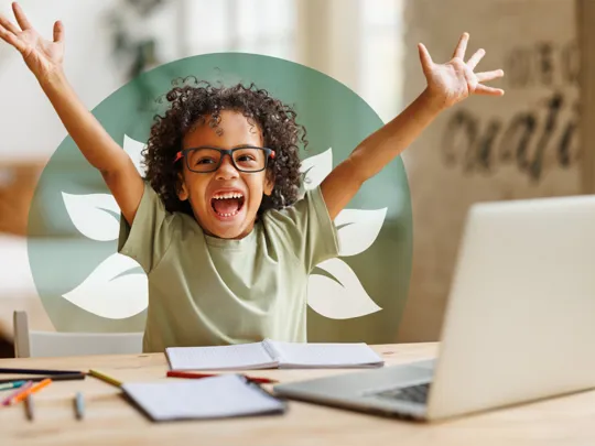 A young student with glasses, smiling excitedly with arms raised, sitting at a desk with a laptop and open notebook.