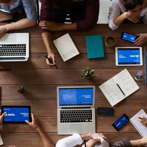 An overhead shot of a group of people sitting around a large wooden table. They are using various devices, including laptops, tablets, and smartphones, and have notebooks and coffee mugs on the table.