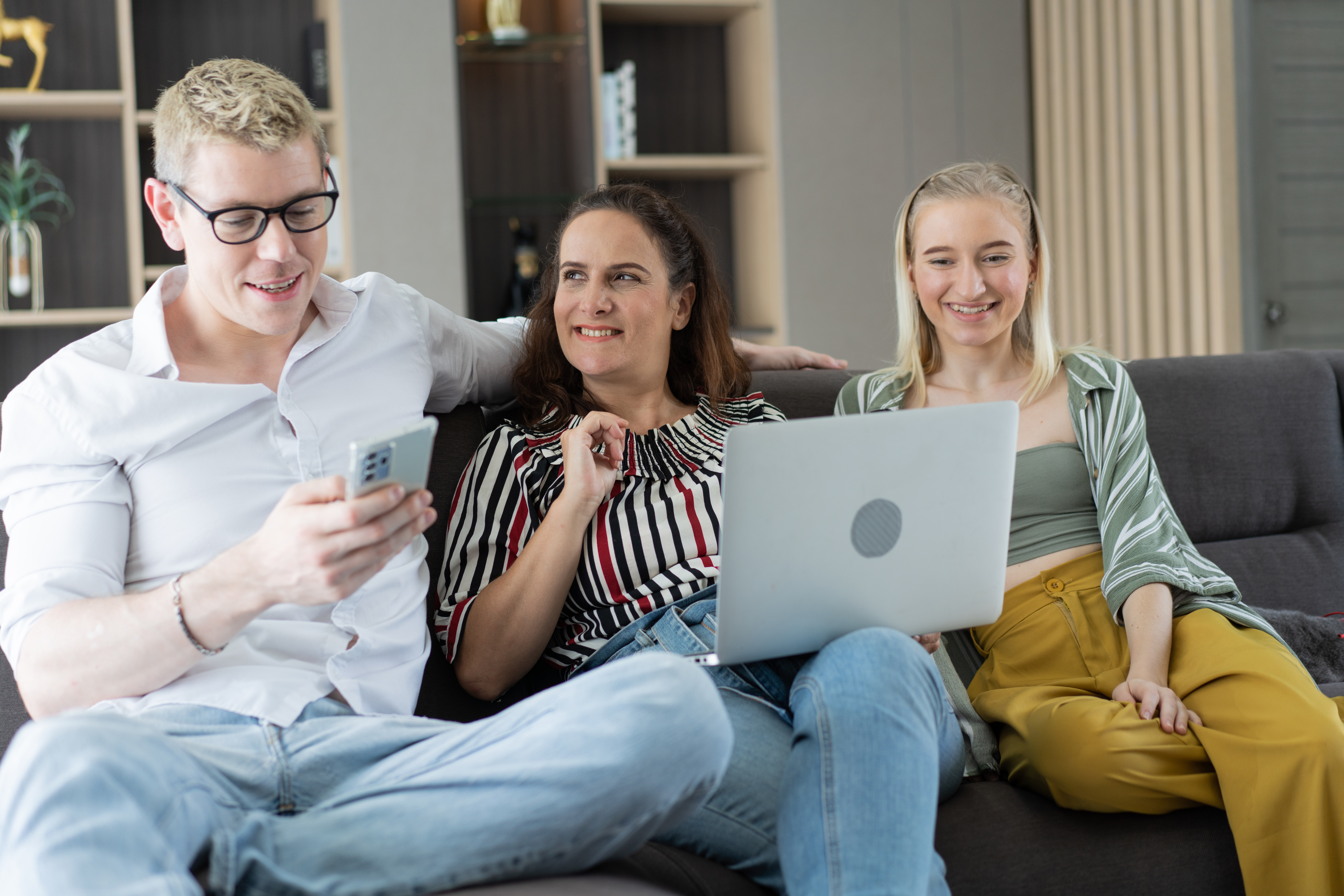 Three people sitting on a sofa smiling while looking at a laptop and phone together