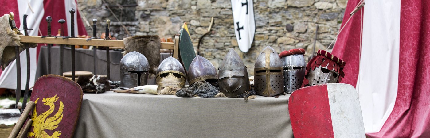 Table displaying medieval helmets, shields and swords in front of a stone wall and tents