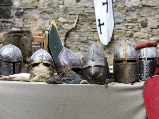 Table displaying medieval helmets, shields and swords in front of a stone wall and tents