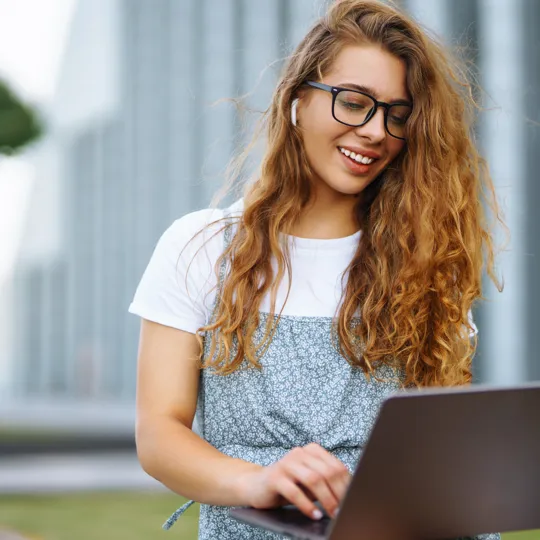 A happy young woman with long, curly auburn hair, wearing glasses and a patterned dress, works on a laptop outdoors. She has wireless earbuds in her ears and is smiling while looking down at the screen, with a modern building in the blurred background.