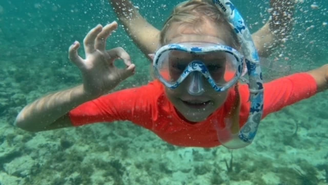 A child wearing a snorkel mask and orange top underwater, giving an 