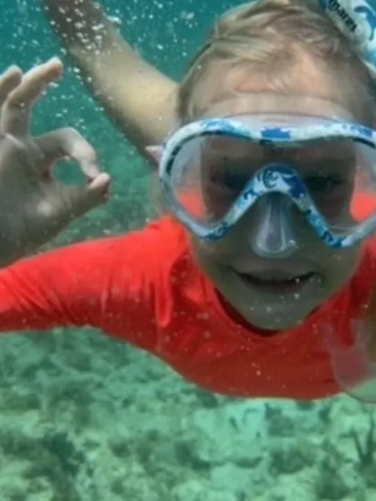 A child wearing a snorkel mask and orange top underwater, giving an