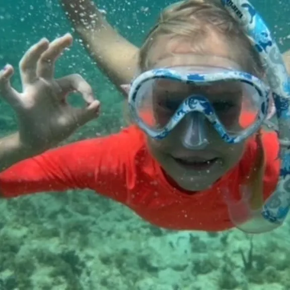 A child wearing a snorkel mask and orange top underwater, giving an