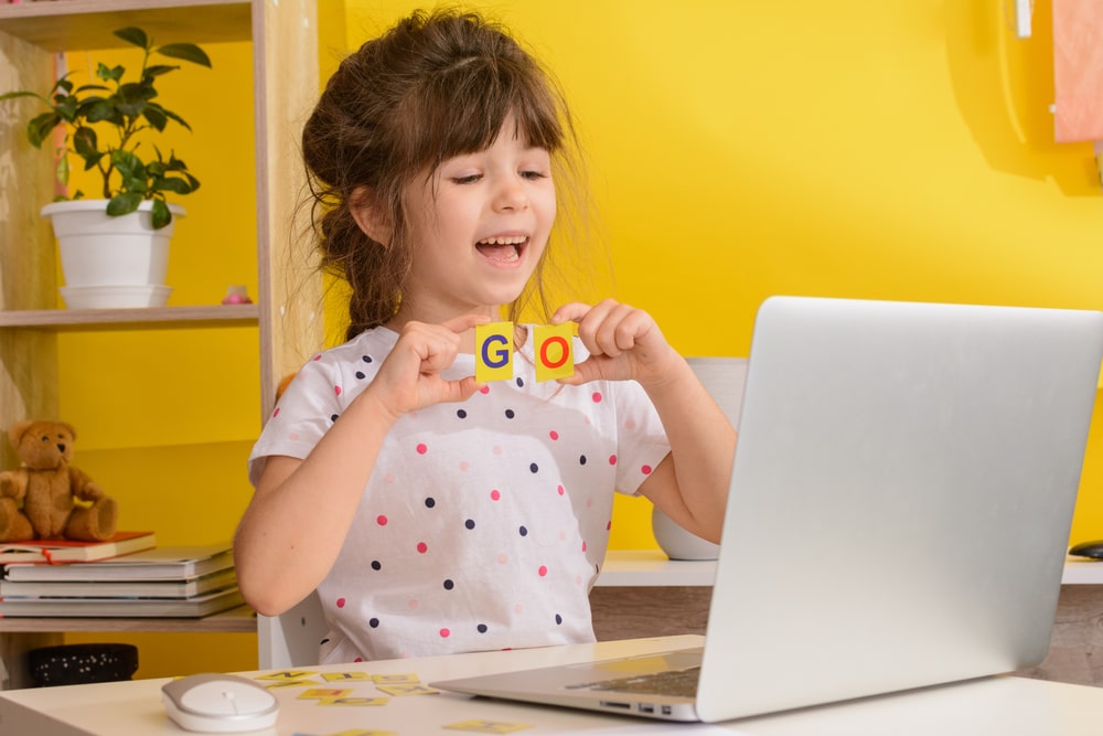 Young girl smiling during an online english session, holding up letter blocks in front of a laptop