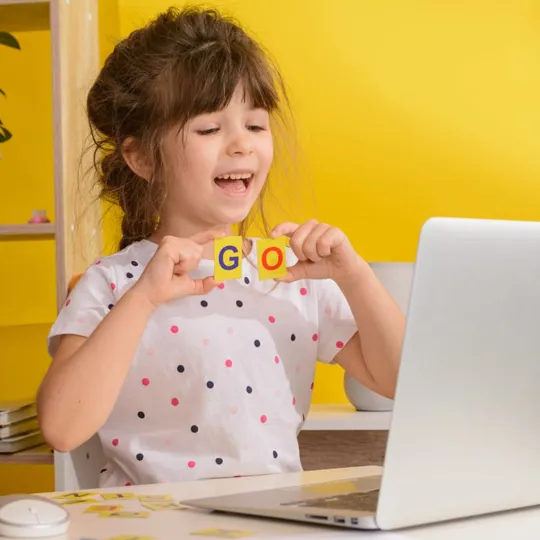 Young girl smiling during an online english session, holding up letter blocks in front of a laptop