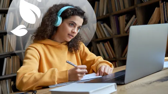A student with curly hair wearing headphones, writing in a notebook while looking at a laptop, with a blurred bookshelf background and a green leafy design.