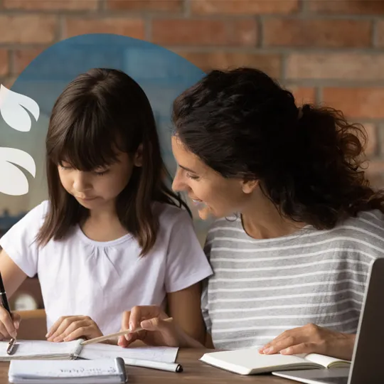 An adult smiling and assisting a young person who is writing in a notebook at a wooden table, with a laptop nearby.