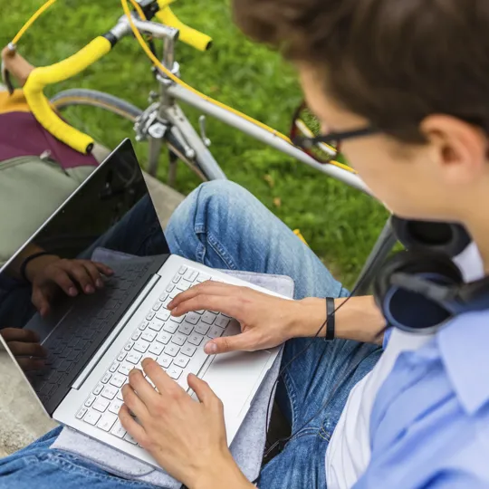 Overhead view of a male student wearing glasses and headphones using a laptop outside, emphasising digital well-being and safeguarding in online education.