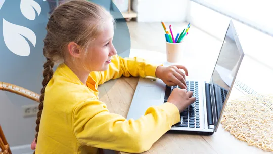 A young student with braided hair, wearing a yellow top, looking at a laptop and typing at a wooden desk.