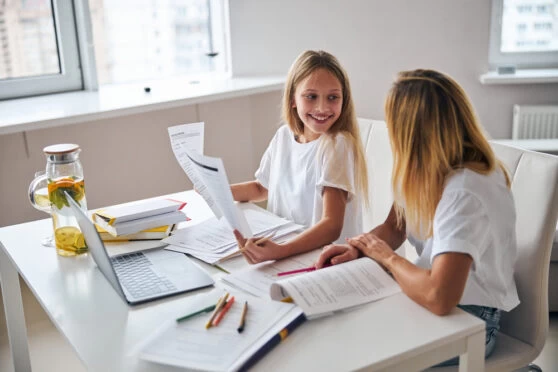 A homeschooling young girl and her mum sitting at a desk talking. The desk is covered with learning materials and papers as well as a laptop and pens. The girl is showing some papers to her mum and smiling.