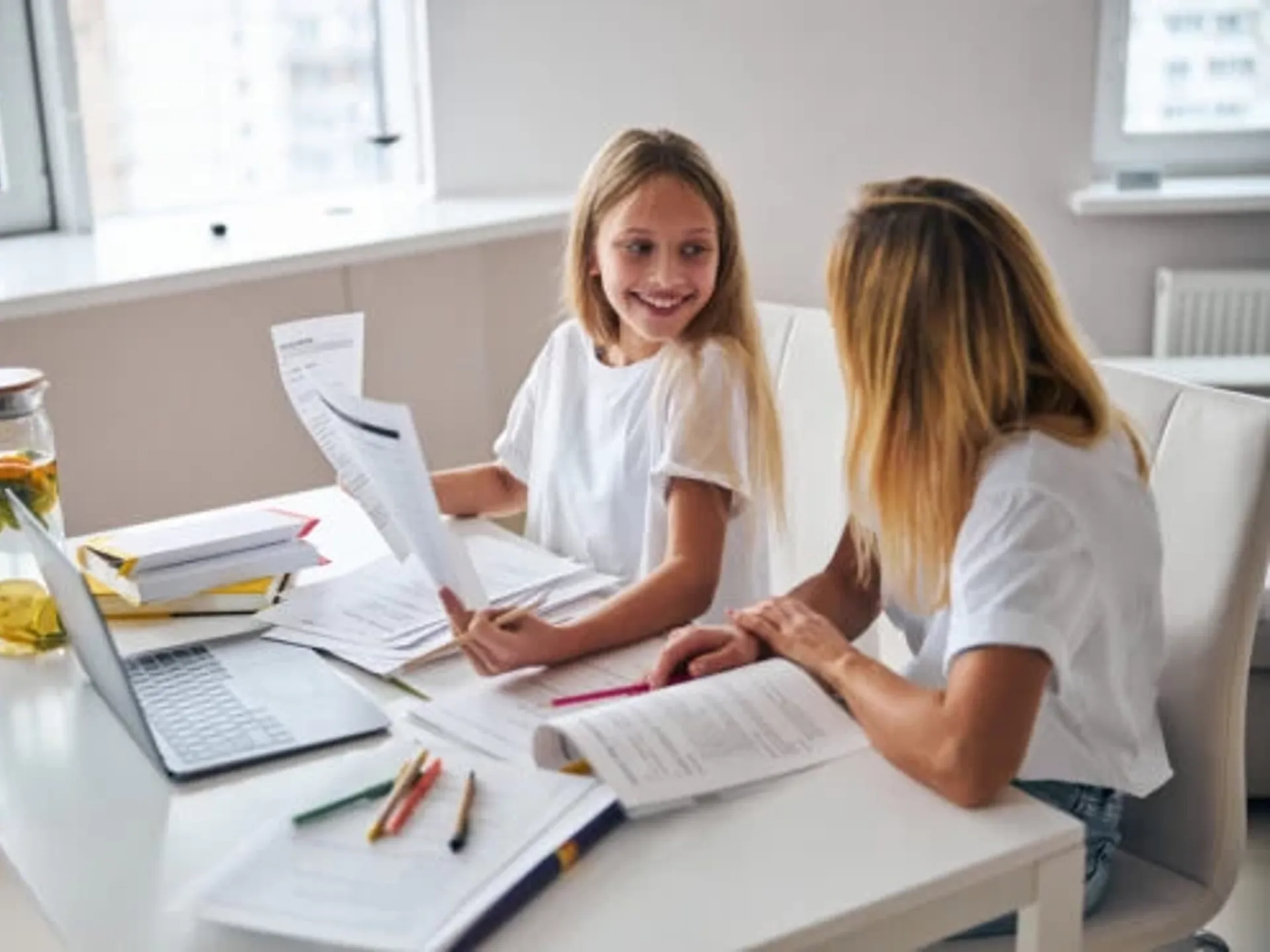 A homeschooling young girl and her mum sitting at a desk talking. The desk is covered with learning materials and papers as well as a laptop and pens. The girl is showing some papers to her mum and smiling.