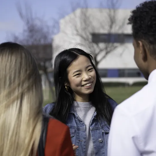 A young woman with long dark hair and a denim jacket smiles while talking to two classmates outdoors on a sunny university campus.