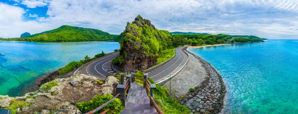 A landscape view of a Mauritian road curving around an outcrop of land over crystal blue sea waters. 