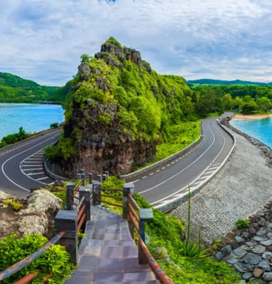A landscape view of a Mauritian road curving around an outcrop of land over crystal blue sea waters.