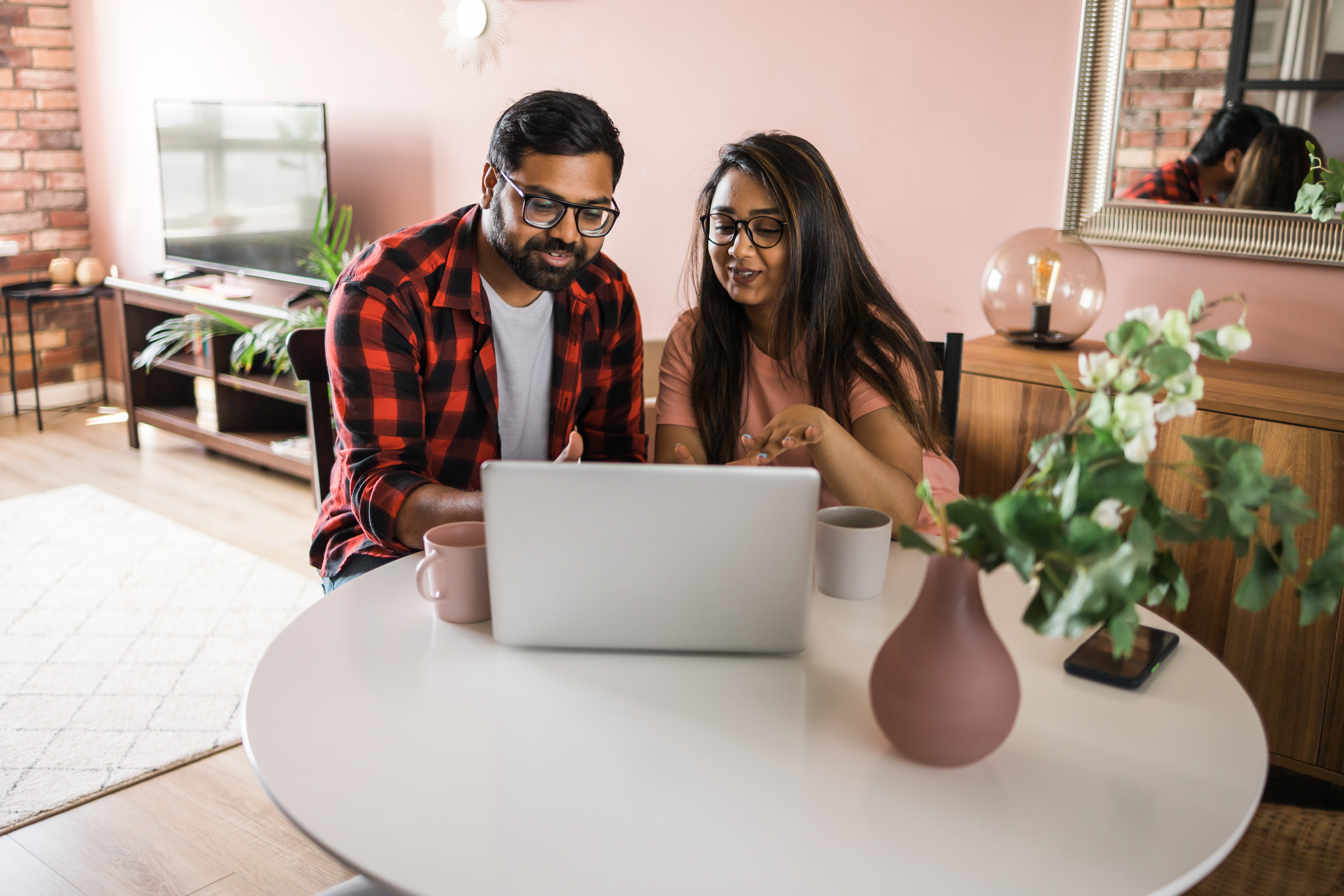Smiling couple seated at a round table looking at a laptop with mugs beside them