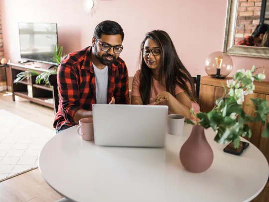 Smiling couple seated at a round table looking at a laptop with mugs beside them